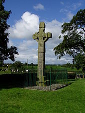 Grande croix en pierre, avec des motifs sculptés complexes, protégée par des balustrades métalliques entourées d'herbe coupée court.  Les arbres sont de chaque côté, les vaches en pleine campagne sont au milieu.