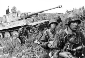A medium tank advancing through a field surrounded by German soldiers.