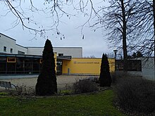 flache Hausfassade: gelber Teil gerade, mit der Aufschrift Hermann-Gocht-Haus, links daneben eine gewölbte Fassade aus Glaselementen. An die genannten Teile schließen graue Elemente an. Im Vordergrund Bänke und Grünbewuchs.