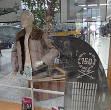 A color photo of a display case, inside with a mannequin doll wearing a fur collar jacket next to an aircraft tail rudder. The rudder shows various markings, on the top the number 150, encircled by a wreath. Below the wreath are two crossed swords and numerous white stripes.