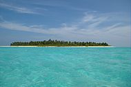 Daytime edge-on view of a small forested island amid a shallow turquoise lagoon. The dark sky blue above is streaked by thin, wispy clouds of various sizes. The lone island is evenly low-slung, no part of it reaching any substantial height whatever.