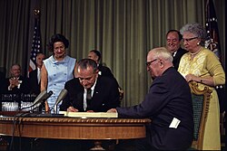 Zwei Männer an einem Schreibtisch mit einem Dokument, das einer unterschreibt, ihre Frauen stehen hinter ihnen