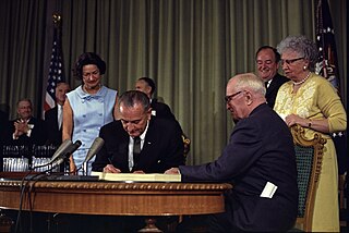 President Lyndon B. Johnson signing the Medicare Bill at the Harry S. Truman Library in Independence, Missouri. Former President Harry S. Truman is seated at the table with President Johnson. The following are in the background (from left to right): Senator Edward V. Long, an unidentified man, Lady Bird Johnson, Senator Mike Mansfield, Vice President Hubert Humphrey, and Bess Truman.