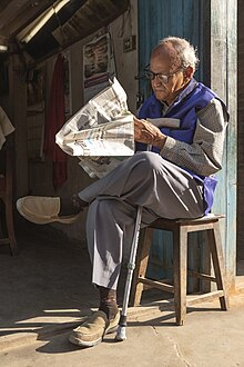 Photograph of a bespectacled person sitting on a stool with their legs crossed reading a newspaper in the morning