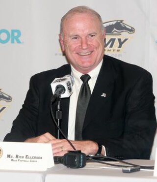 Man in a black suit sitting at press conference table with microphone in front of him