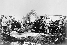 Fotografia di un imponente cannone protetto da sacchi di sabbia e circondato da numerosi cannonieri in divise disparate