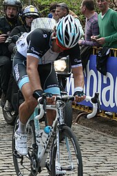 A road racing cyclist wearing a black and white jersey with pale blue trim, riding with his head down. A motorbike follows behind him, and spectators watch on from behind roadside barricades.