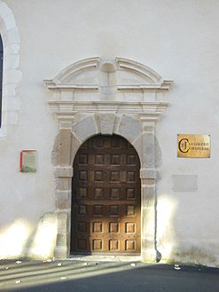 Fotografia de uma porta de madeira com o topo arredondado destacando-se contra uma parede branca.