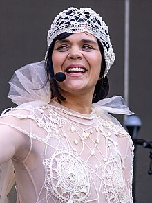 Natasha Khan wearing a frilly, sheer top, with an embroidered cap, squinting and appearing to sing into a wireless microphone while looking right of camera with right arm extended