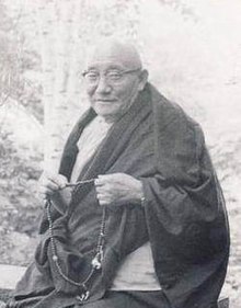 Dezhung Rinpoche III, likely Seattle, date unknown Buddhist scholars - Dezhung Rinpoche III seated on a stool, Chris Wilkinson, student seated at the feet of his lama, wearing Buddhist robes, malas, likely Seattle, Washington, USA (16134833232) (cropped).jpg