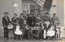 Schwarz-Weiß-Foto von fünfzehn Personen vor einem Fenster.  Die Männer sind als Wehrpflichtige verkleidet und einer von ihnen trägt eine Flagge.