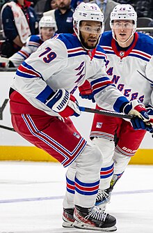 A young black man in a New York Rangers jersey holding a hockey stick in his left hand, looking down and to the side.