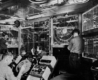 Men stand at various terminals scattered around a cramped room