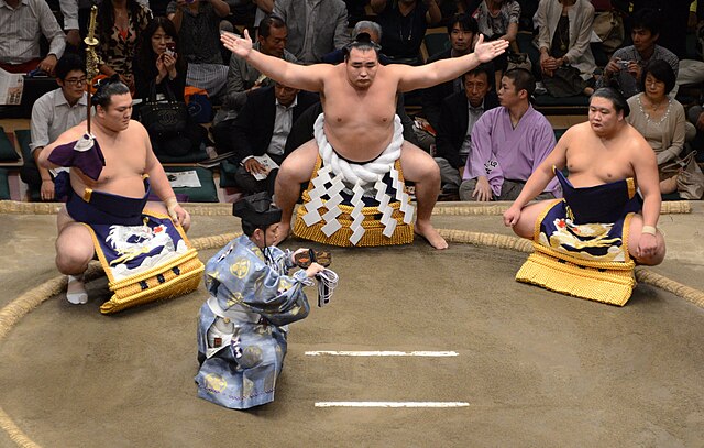Yokozuna Kakuryū Rikisaburō (center) performing the ring-entering ceremony while flanked by a sword bearer on the left and dew sweeper on the right