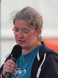 A woman with glasses and gray hair holds a microphone while speaking at an indoor event.