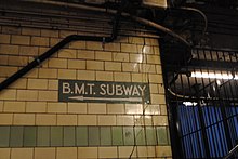 Cremefarben geflieste Bahnhofswand mit einem Wegweiser aus einem braunen Keramik-Mosaik mit der Inschrift „B.M.T. Subway“ und einem Richtungspfeil.