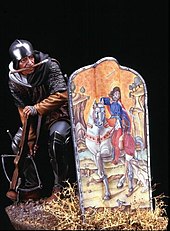 A model of a medieval crossbowman drawing his bow behind his pavise. A hook on the end of a strap on his belt engages the bowstring. Holding the crossbow down by putting his foot through the stirrup, he draws the bow by straightening his legs Balestriere1.jpg