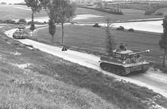 Quatro tanques descem por uma estrada arborizada em campo aberto.