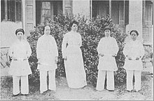 Five women standing in a row outdoors. The women at the center is the tallest, and is a white woman wearing a white dress. The rest are Chinese women, wearing trousers and long smocks.
