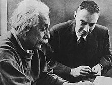 Einstein écrit à un bureau.  Oppenheimer est assis à côté de lui, le regardant.