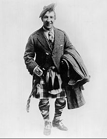 Black and white image of Harry Lauder in a kilt, tartan hose and tie, tweed jacket, and a Balmoral bonnet