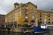 Umgebautes Docklager in Buff Brick mit einer markanten Brüstung.  In der Brüstung über der Säule der Ladetüren ist eine Lücke sichtbar, in der eine Lucarne entfernt wurde.  Die Türen haben auch klappbare Eisenklappen, die durch Ketten als Ladeverlängerungen festgehalten werden.