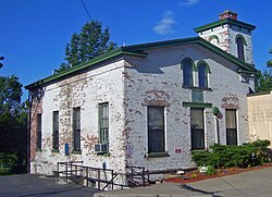 Un petit bâtiment en brique avec de la peinture blanche s'écaillant par endroits, des garnitures vertes, un toit à pignon en pente douce et une tour basse derrière le coin droit.