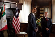 Felipe Calderón standing on the left shakes hands with Obama on the right. Both presidents are smiling towards the camera, posed in front of the national flags.