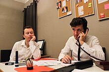 Deux personnages autour d'un bureau dans un bureau ; l'un, affalé, pensif, l'autre, au téléphone, sérieux.