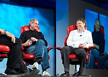 Two middle-aged men shown full length, sitting in red leather chairs and smiling at each other
