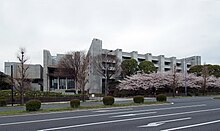 Bâtiment moderne en pierre grise, une double rangée d'arbres, la plus proche en fleur, bordant le coté de la rue que longe le bâtiment.