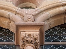 Fotografie color care prezintă un capitel de fereastră sculptat cu frunze de struguri și depășit de un cap uman și un oculus.  Deschiderile au panouri de vitralii în formă de diamant.