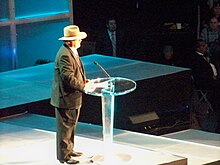 Homem na frente de uma mesa e um microfone.