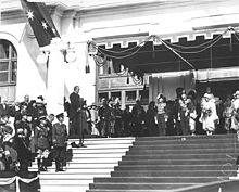 Photographie d'un homme en costume derrière un pupitre au sommet d'un escalier devant l'entrée d'un bâtiment. Des dizaines de personnes en uniformes militaires ou en costumes assistent au discours.