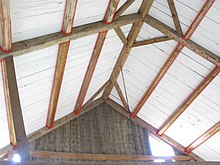 A view of a roof using common purlin framing. The purlins are marked in red. This view is from the inside of the building, below the roof. The rafters are the beams of wood angled upward from the ground. They meet at the top of the gable at a ridge beam, which has extra bracing to attach it to the rafters. The purlins are the large beams perpendicular to the rafters; from this shot, it appears that there are three purlins on either side of the roof. The sheathing boards are sometimes called the roof deck and are painted white. Common Purlin Highlighted.jpeg