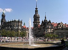 Barevná fotografie s pohledem na tryskající fontánu, za níž je vpravo rezidenční zámek a vlevo věž Hofkirche