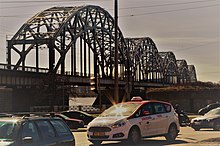 Seitliche Farbfotografie einer Eisenbrücke mit fünf Rundbögen, die über die unten liegende Fahrbahn führen. Im Vordergrund fahren Autos auf zwei sich treffenden Straßen. Zwischen den Unterbauten ist links der Fluss zu sehen.