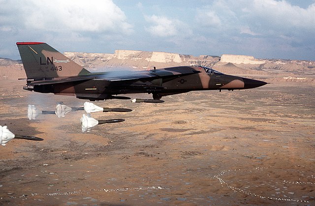 Vue rapprochée d'un F-111 en train de larguer quelques bombes freinées. L'avion vole à vitesse modérée à basse altitude, au-dessus du désert.