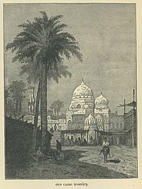 A man on a donkey walks past a palm tree, with a mosque and market behind him.