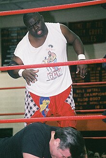 Un luchador negro con una camiseta blanca frente a su oponente en el suelo.