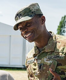 Candid photo of a young black man; he is wearing camouflage fatigues, looking down and to the camera's left, and smiling.