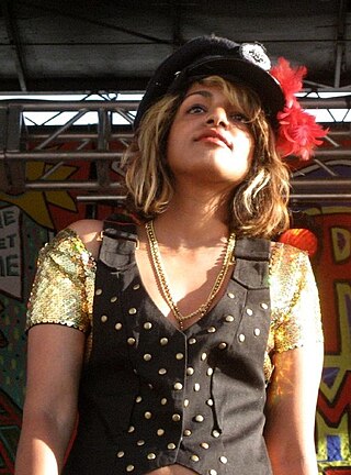 M.I.A. standing in a golden t-shirt, coat and a hat rimmed with flowers.