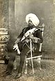 A photograph of a man seated on an ornate chair beside a similarly ornate vase. He is wearing a turban, full-dress military uniform and the sash, collar and star of the Order of the Star of India; he is holding in his left hand a sword and in his right hand two dress gloves.