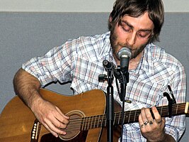 close-up of Mike Kinsella wearing a light checkered shirt, playing an acoustic guitar and singing into a microphone