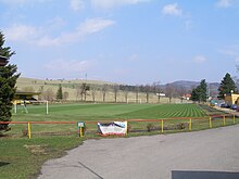 Zatravněné fotbalové hřiště místního fotbalového stadionu. Hřiště je obehnáno kovovým zábradlím. Před ním je zpevněná asfaltová plocha. V pozadí je Mořkovský vrch, na kterém je základnová stanice mobilního operátora a vede tudy vysoké elektrické napětí.