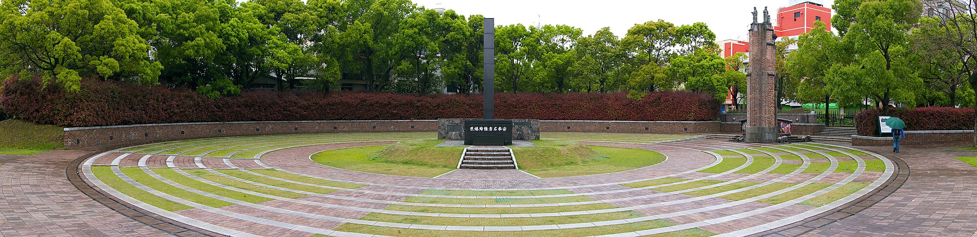A rectangular column rises above a dark stone base with Japanese writing on it. It sits atop a grass mound which is surrounded by alternating circles of stone path and grass. There is a wall around the whole monument, and bushes beyond.