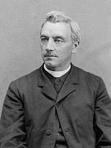 Black and white photo of an older man wearing black with a priest's colar and facing right.