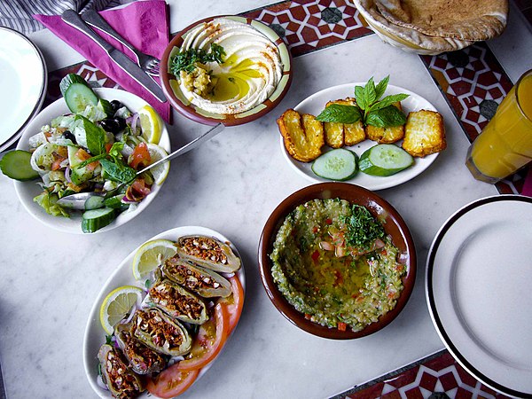 A typical Syrian meal beginning at lower left of center, and continuing clockwise: makdous, syrian salad, hummus, haloumi and baba ganouj, with pita bread partially visible at upper right corner of photo.