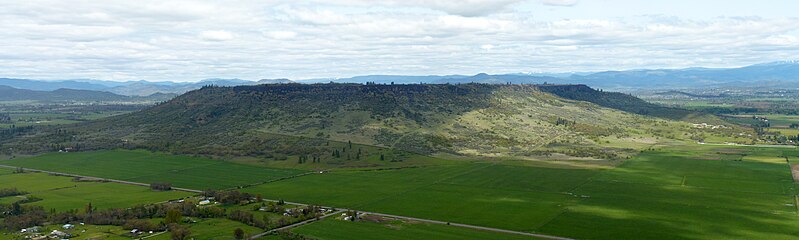 A flat topped mesa surrouned by cliffs and tree-covered slopes with farmland below
