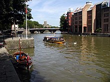 Een gele watertaxi op het water tussen stenen kades.  De verre oever heeft grote gebouwen en in de verte is een brug met drie bogen.